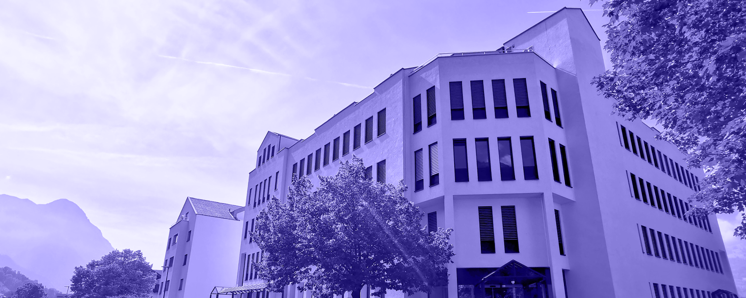 A contemporary white office building with large windows and shutters, set against a scenic backdrop of mountains and trees on a clear day. The building is surrounded by greenery, with a bright, slightly purple-tinted sky above.